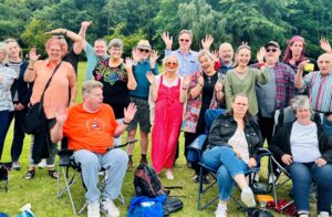 A group of people who use the Friends in Need Bracknell service, smiling and waving. Three sitting at the front and the rest standing behind. They are in an open, green space with trees behind them.