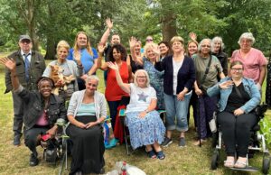 A group of people who use the Friends in Need Slough service, smiling and waving. Three sitting at the front, one person crouching at the front and the rest standing behind. They are in a wood with trees around them.