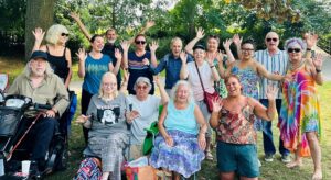 A group of people who use the Friends in Need Windsor, Ascot and Maidenhead service, smiling and waving. Three sitting at the front. two are kneeling at the front and the rest standing behind. They are in a green space with trees around them.