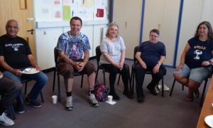 A group of people smiling and sitting in chairs in a semi-circle. They are in Bucks Mind's High Wycombe premises and are members of the Mind Gap Peer Support service.