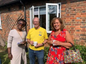 Three volunteers, a man and two women, standing outside in front of an open window. They are smiling and holding coffee cups.