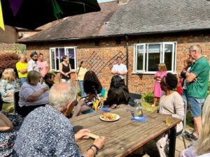 A group of people, Bucks Mind's volunteers, standing and sitting in the Sun House garden while enjoying refreshments and hearing a short speech from Stephen Robinson, Deputy Chief Executive at Buckinghamshire Mind/