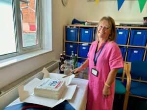 A woman in a pink dress is smiling while cutting a large cake with Bucks Mind's 110th anniversary logo featured.